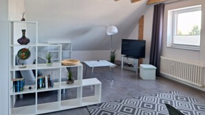 A modern living room with a sloped ceiling, built-in white shelving, a flat-screen TV, a white table, and a patterned rug.