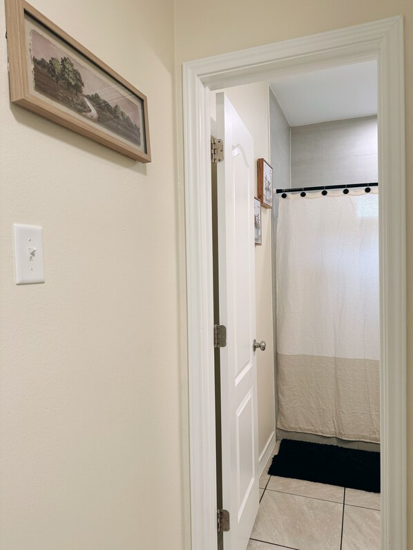 A bathroom with a shower curtain, a door with a handle, a light switch, and a framed picture on the wall.
