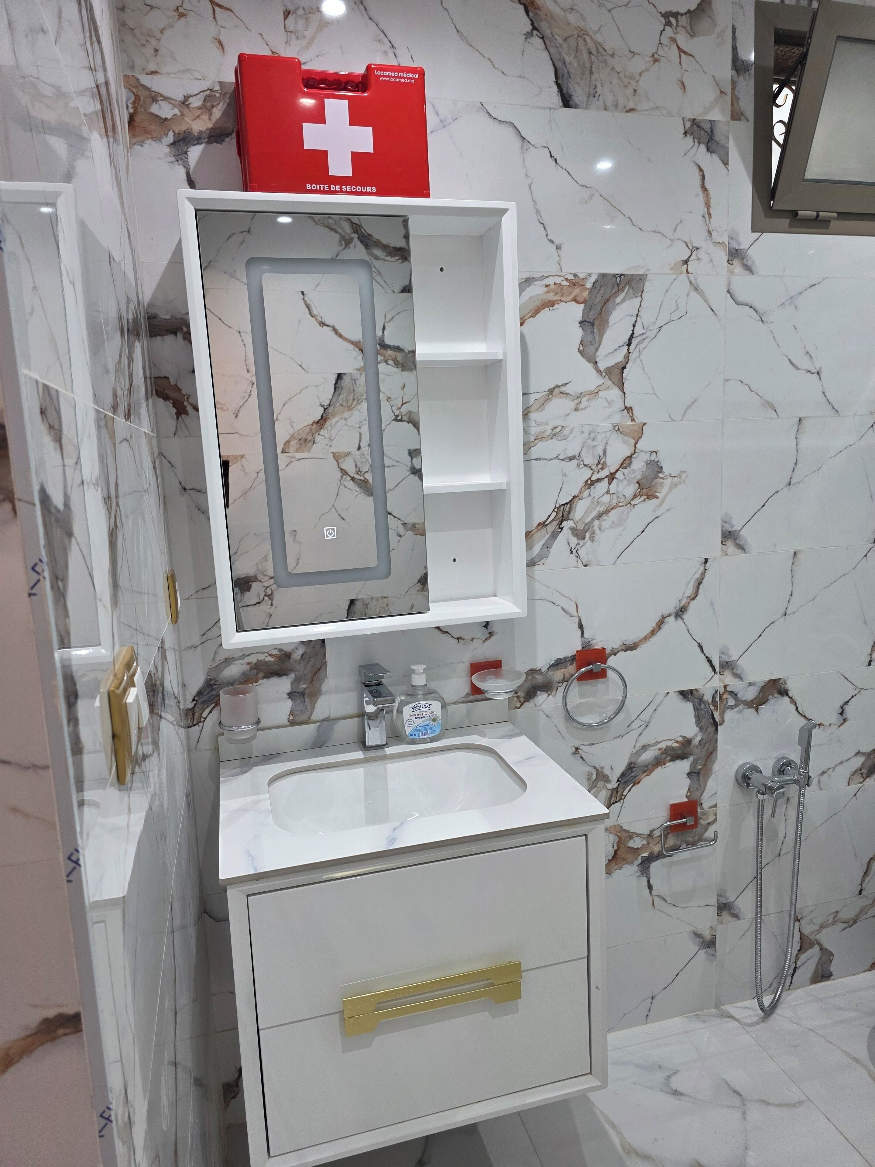 A bathroom with a marble backsplash, a white sink, a mirror with shelves, a red first aid kit, and a shower area.