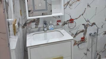 A bathroom with a marble backsplash, a white sink, a mirror with shelves, a red first aid kit, and a shower area.