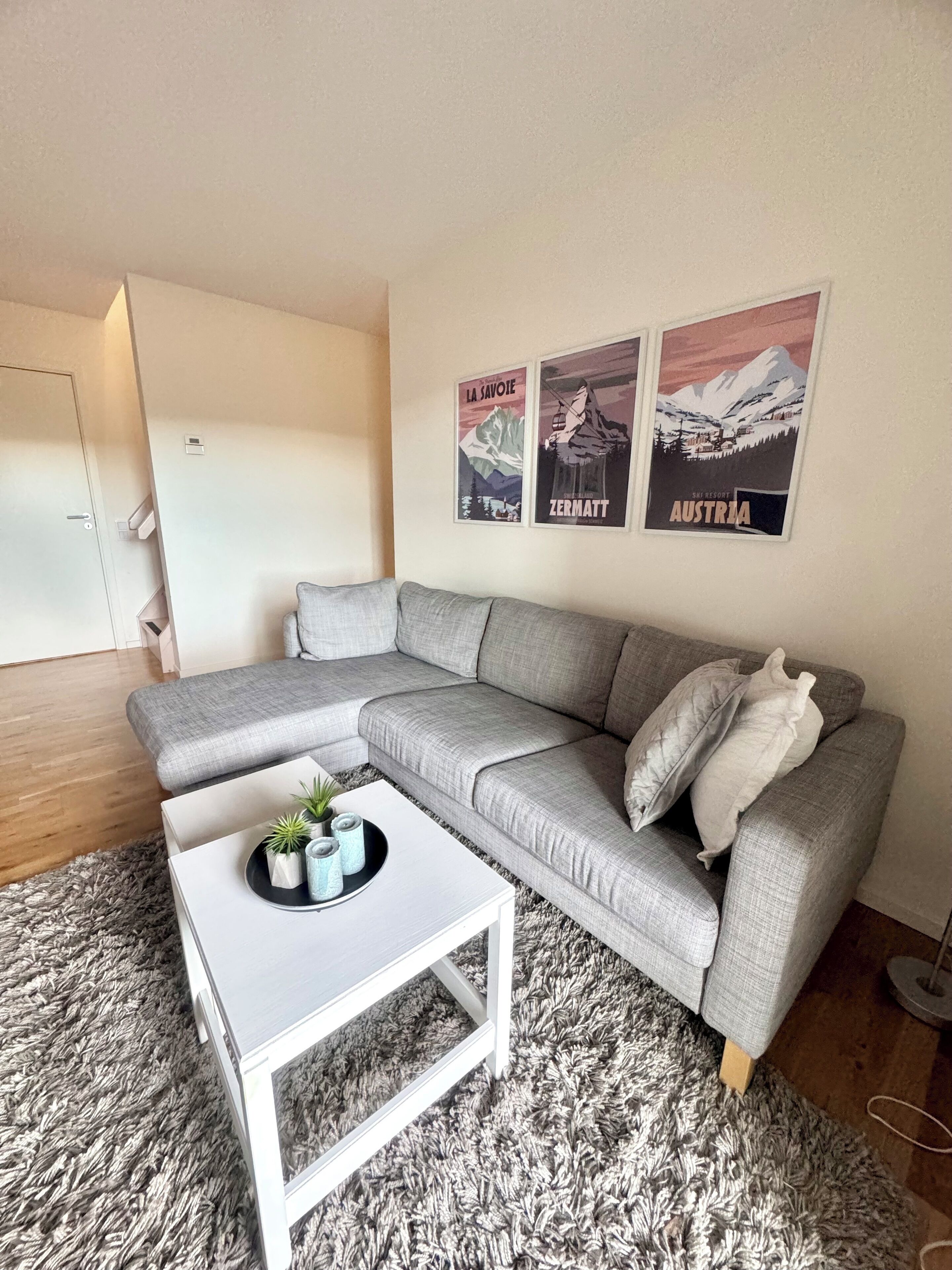 Ein modernes Wohnzimmer mit einem grauen Ecksofa, einem weißen Couchtisch mit Dekoration und zwei gerahmten Postern an der Wand.