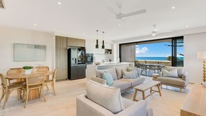 Een moderne woonkamer met een grote bank, een eethoek met rieten stoelen, een keuken met een inbouwkoelkast, en een balkon met uitzicht op de oceaan.