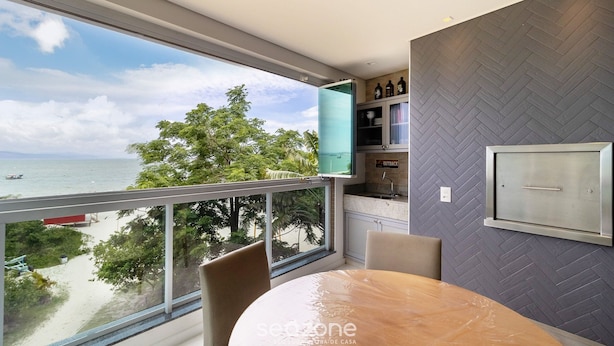 A modern room with a large window overlooking a beach, a round table, and a herringbone-patterned wall. - Apartment Front to the Sea With Barbecue Vdr0202 (Florianópolis)