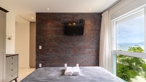A bedroom with a brick wall, a flat-screen TV, a bed with white linens, and a view of the sea through a large window. - Apartment Front to the Sea With Barbecue Vdr0202 (Florianópolis)