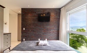 A bedroom with a brick wall, a flat-screen TV, a bed with white linens, and a view of the sea through a large window.