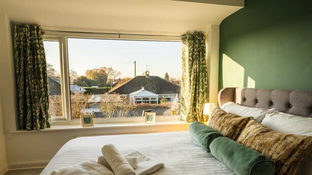 A bedroom with a large window, a bed with white linens, and a view of houses outside. - A  cosy place to stay near Manchester Airport (Heald Green)