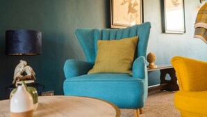 A teal armchair with a mustard pillow, a yellow armchair, a round wooden table, and a lamp with a dark shade. - A  cosy place to stay near Manchester Airport (Heald Green)