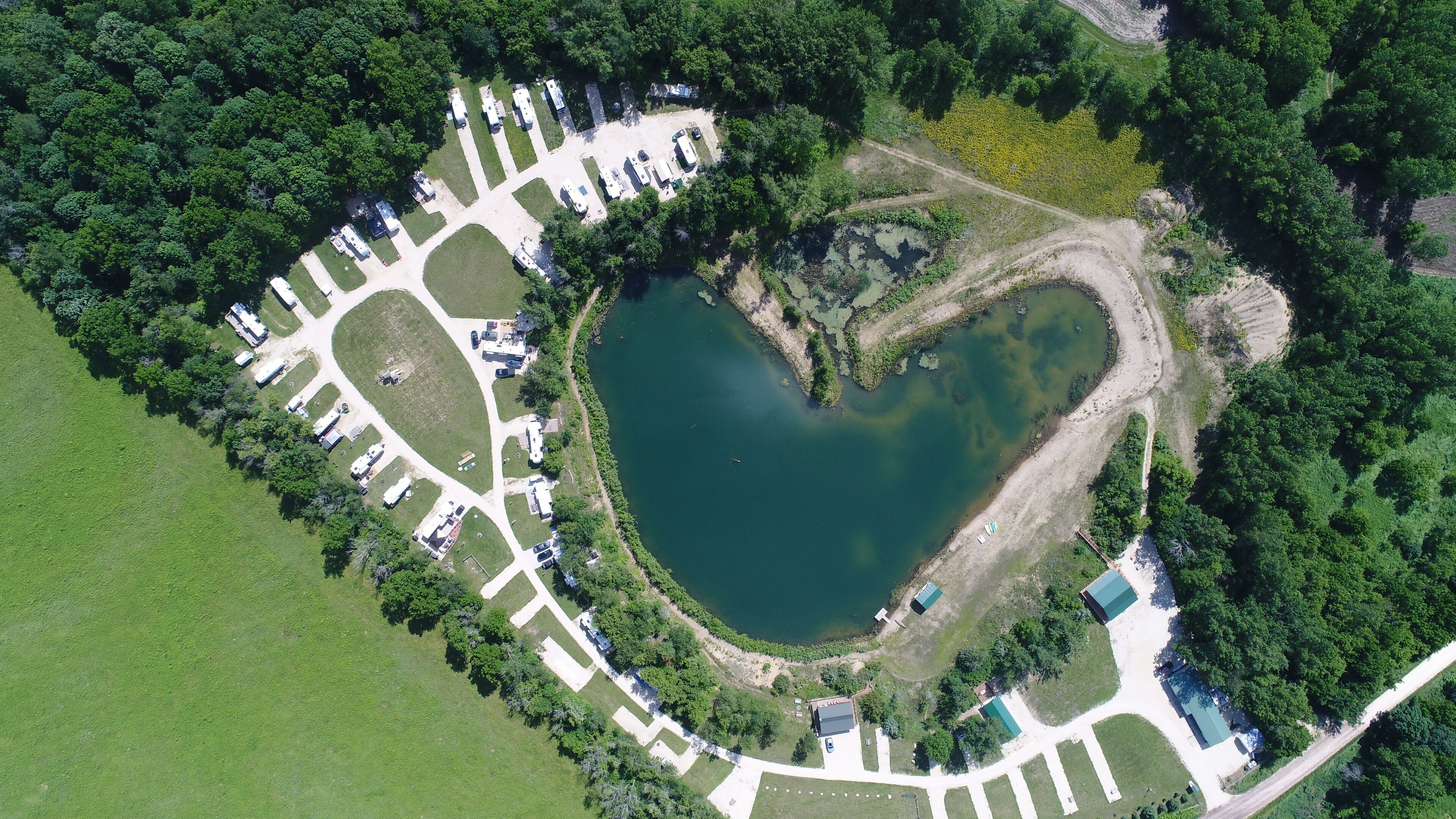 Une vue aérienne d’un terrain de camping où des véhicules récréatifs sont stationnés dans une zone en forme de cœur, entourée de verdure et d’un lac.