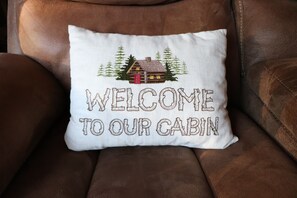 A white pillow with a cabin illustration and the text "WELCOME TO OUR CABIN" on a brown leather chair.