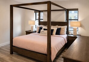 Una cama con dosel de madera, con un edredón rosa y varios almohadones, incluyendo uno con texto. Hay dos mesitas de noche con lámparas, y un banco de madera al pie de la cama.