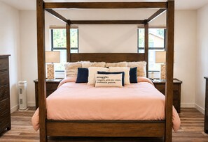 Una cama con dosel y cuatro columnas, una mesita de noche con lámpara y un bulto decorativo en otra mesita de noche.