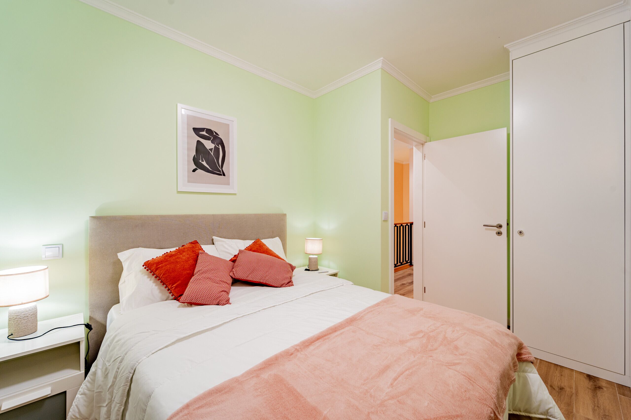 A bedroom with a bed, pillows, a nightstand, a lamp, a framed picture on the wall, and a wardrobe.