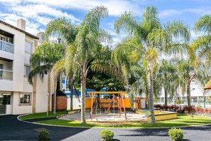 A playground with swings and a yellow play structure, surrounded by palm trees and a building with a tiled facade. - Pleasant Apartment 110m From Ingleses Beach Vdg114 (Florianópolis)