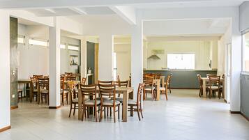 A spacious dining area with multiple wooden tables and chairs, a kitchen in the background, and a high ceiling with exposed beams.