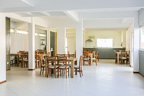 Uma ampla área de refeições com várias mesas e cadeiras de madeira, uma cozinha ao fundo e um teto alto com vigas aparentes.