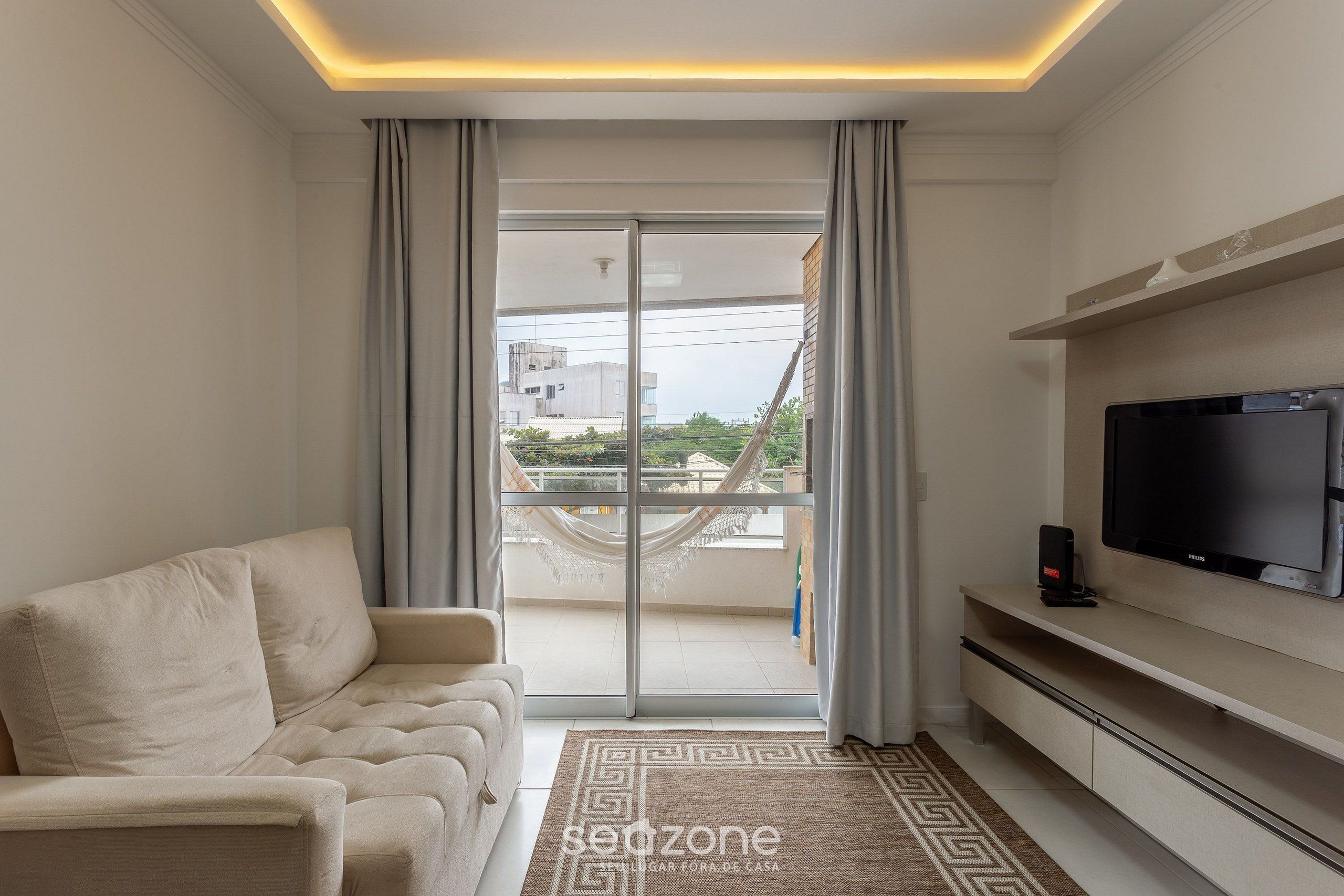 A modern living room with a sofa, a flat-screen TV, and a view of a balcony with a hammock.
