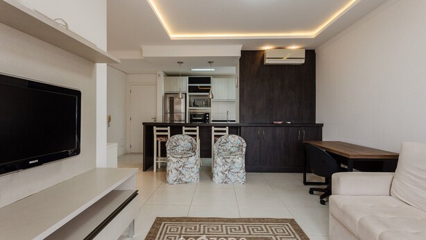 A modern living room with a flat-screen TV, a sofa, two patterned chairs, a wooden table, and a kitchen area with dark cabinets and a refrigerator. - Apt W/bbq, 850m From Ingleses Beach Vde101 (Florianópolis)