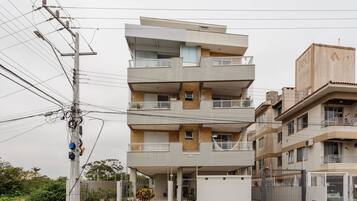 Um edifício residencial de vários andares com varandas, entrada com portão e postes de eletricidade visíveis.