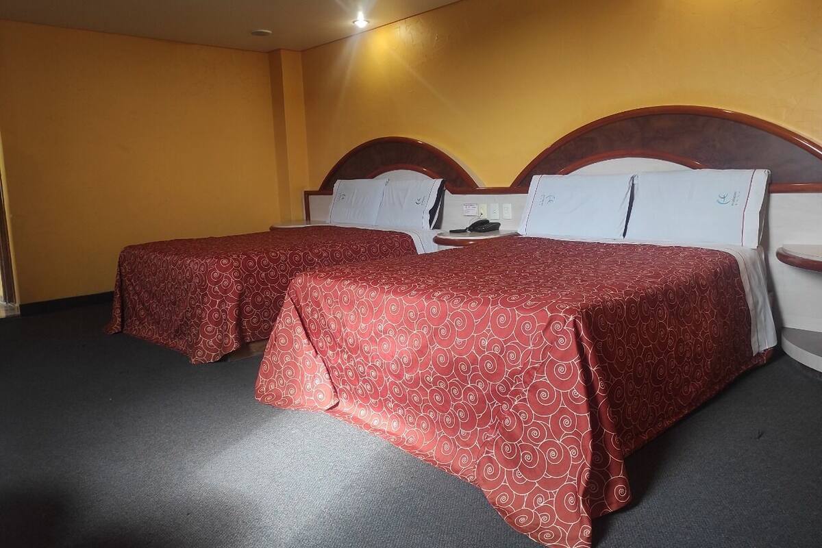 A hotel room with two beds, each with a red patterned bedspread and white pillows.