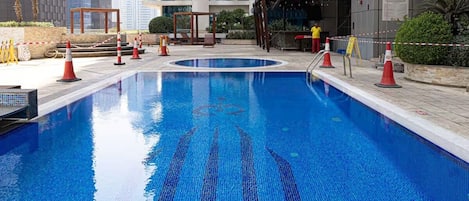 A swimming pool with tiled blue mosaic patterns, surrounded by a modern building with balconies and a covered area with seating.