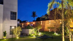 A modern building with a flat roof, a small garden area with a lawn, a palm tree, and a lounge chair. - Confraria Da Prata Flats (Pirenópolis)