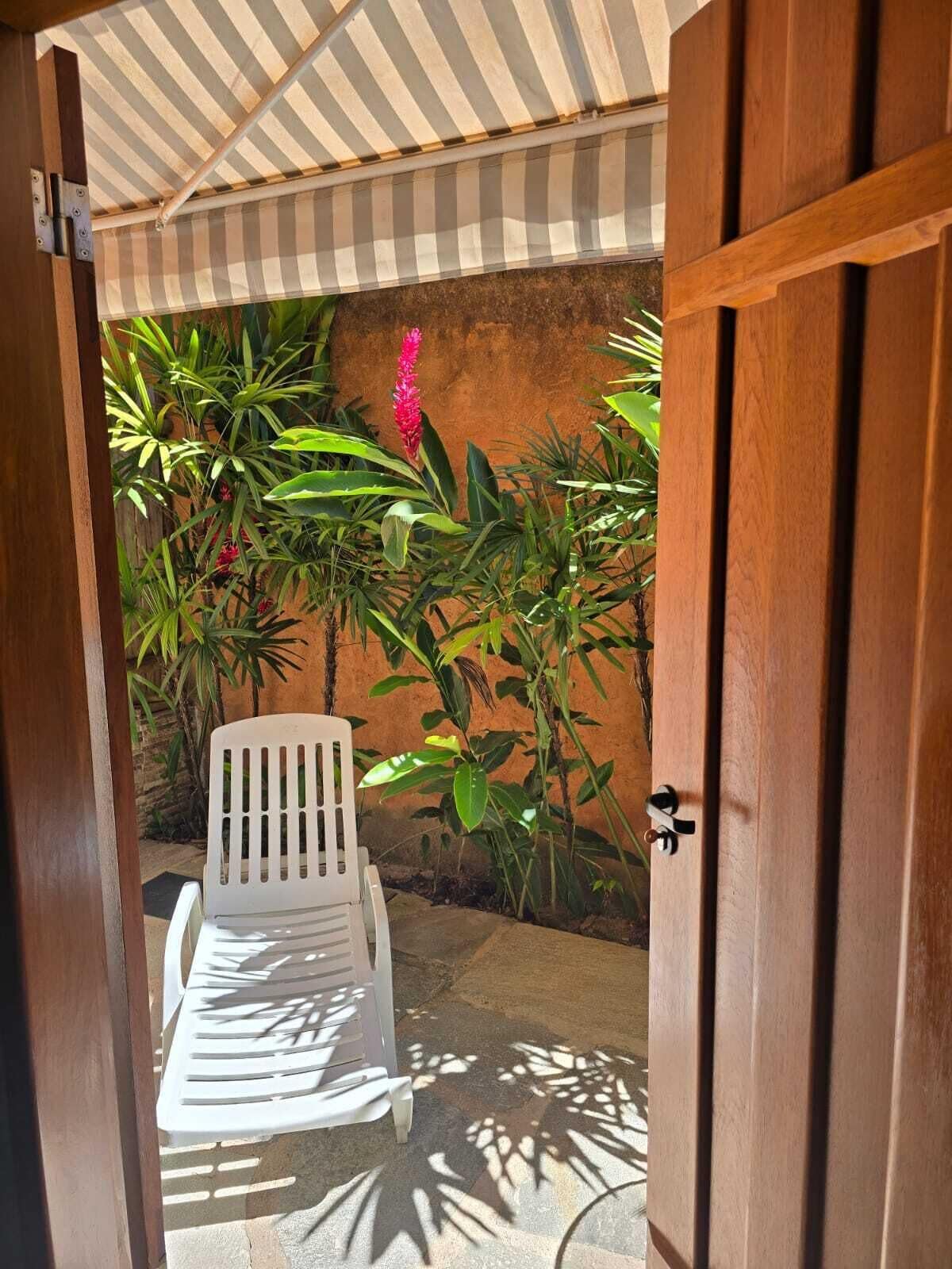 Een open houten deur die leidt naar een overdekt patio met een witte plastic stoel en verschillende tropische planten.
