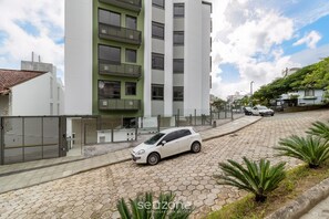 A modern building with a white facade and a green balcony, a white car parked in front, and a paved street with plants. - Apartments Just a few Minutes From Ufsc, Condo With Laundry Facilities (Florianópolis)
