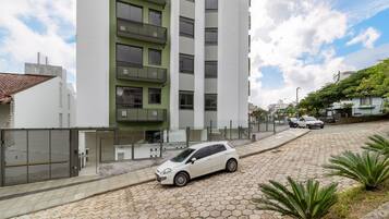A modern building with a white facade and a green balcony, a white car parked in front, and a paved street with plants.