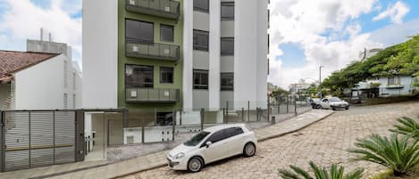 A modern building with a white facade and a green balcony, a white car parked in front, and a paved street with plants.