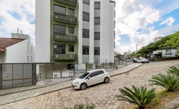 A modern building with a white facade and a green balcony, a white car parked in front, and a paved street with plants. - Apartments Just a few Minutes From Ufsc, Condo With Laundry Facilities (Florianópolis)