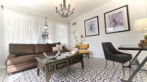 Una sala moderna con un sofá marrón, una mesa de centro de vidrio, una silla negra y una mesa de comedor. Hay dos obras de arte enmarcadas en la pared y una araña sobre el sofá.