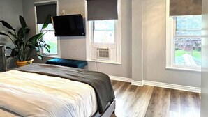 Un dormitorio con una cama, un televisor montado en la pared y dos ventanas con persianas.