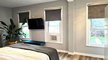 A bedroom with a bed, a TV mounted on the wall, and two windows with blinds.