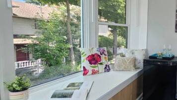 Una habitación con un asiento junto a la ventana, una cesta de frutas y una pequeña mesa con botellas y un cuaderno.