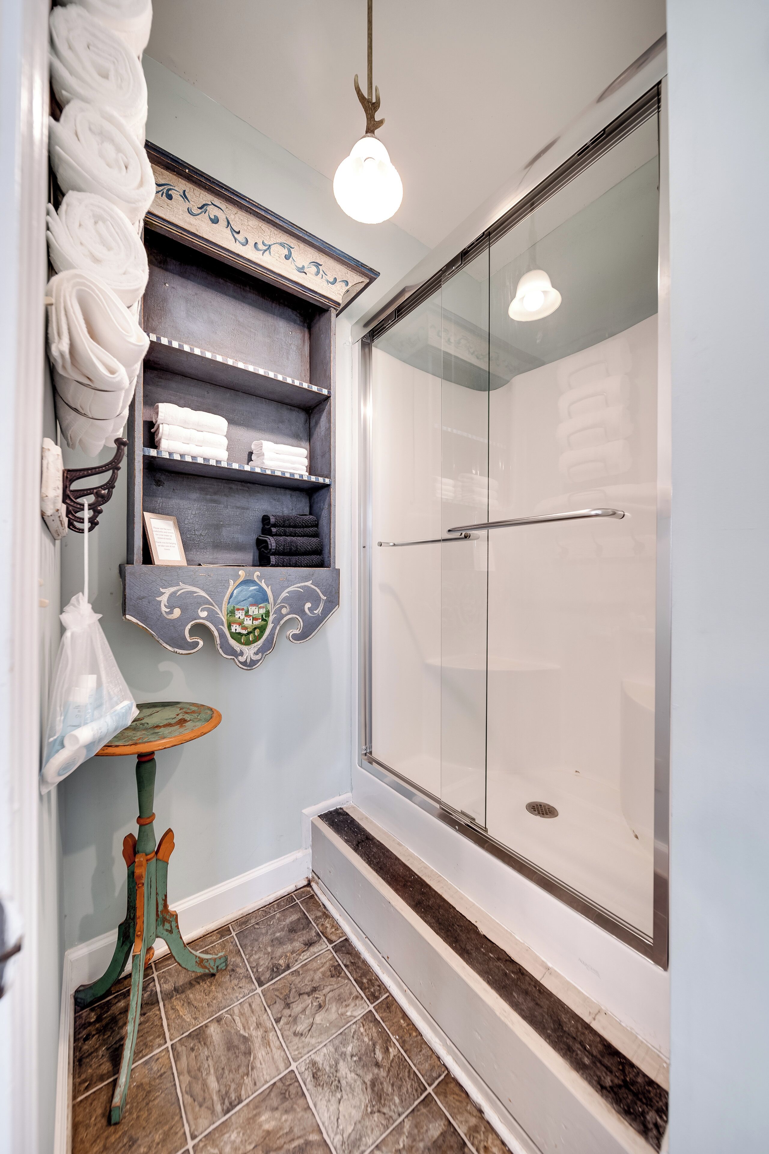 A bathroom with a glass shower, a towel rack with neatly folded towels, a small table with a decorative item, and a shelf with folded towels.