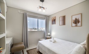 Een hotelkamer met een bed, een bureau, een stoel, een kleine tafel met een lamp en uitzicht op de stad door een groot raam.