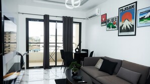 A modern living room with a sofa, a coffee table, and a TV. There is a balcony with a table and chairs, and a city view.