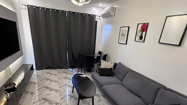 A modern living room with a grey sofa, a black ottoman, a marble floor, a TV, and a dining area with a table and chairs.