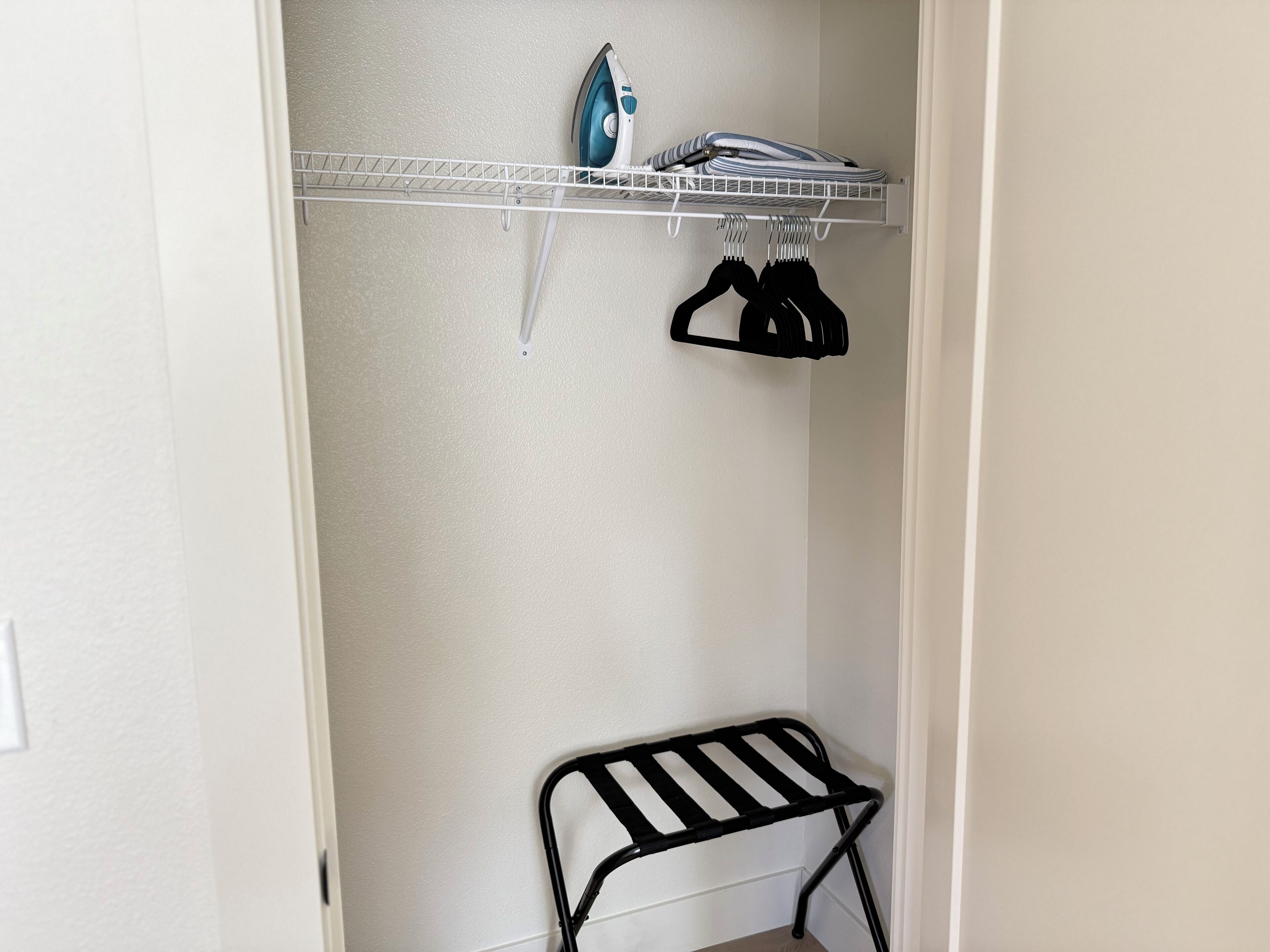 A laundry area with a shelf holding an iron and folded clothes, a drying rack, and a chair.
