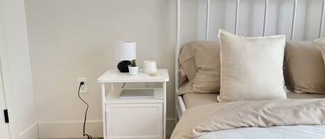 A neatly arranged bedroom with a white metal bed frame, a bedside table with a lamp, a small potted plant, and a bedside table with a notebook and pen.