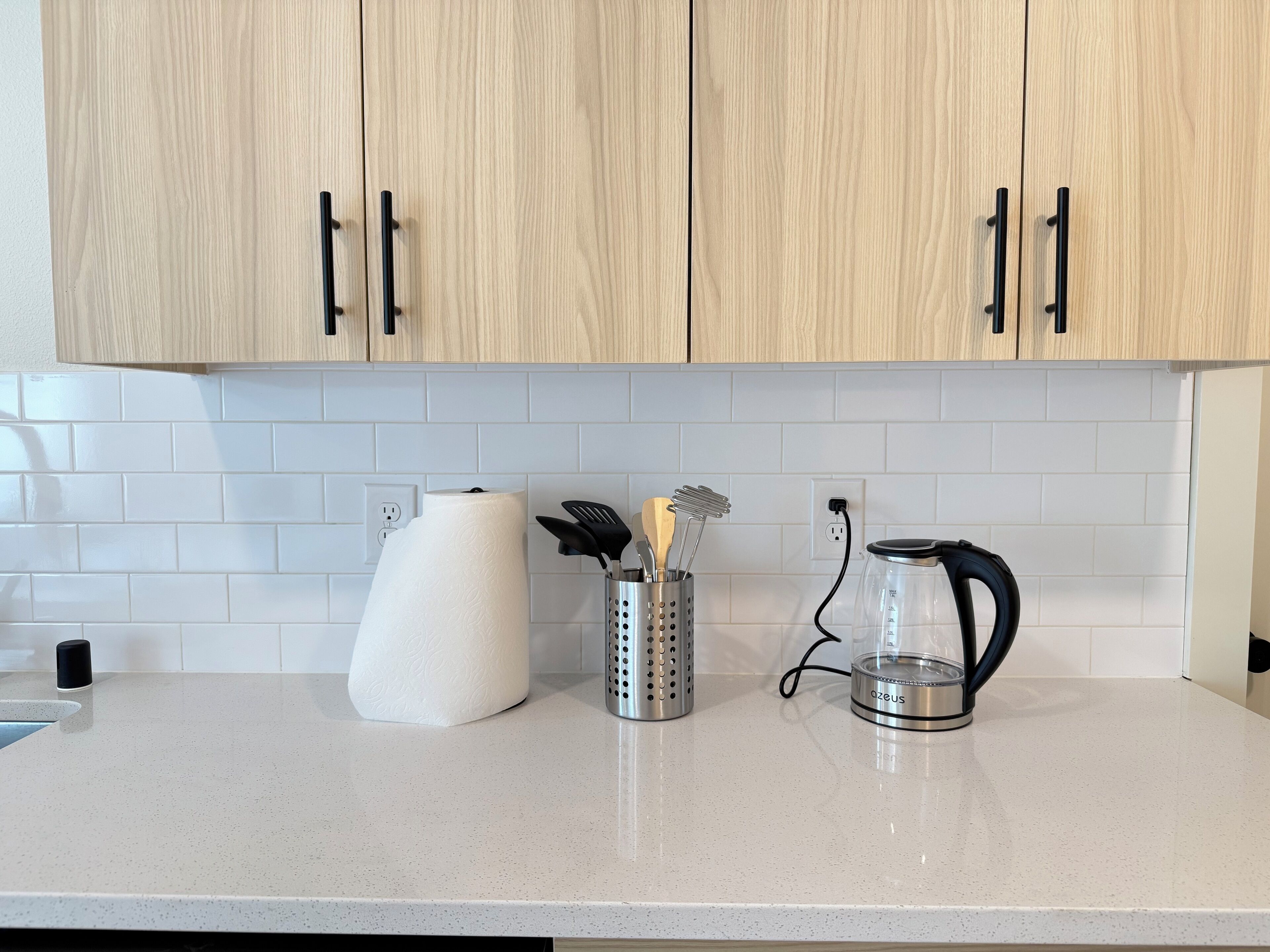 A kitchen with light wood cabinets, white subway tiles, a paper towel holder, a glass kettle, and a utensil holder.