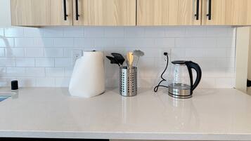 A kitchen with light wood cabinets, white subway tiles, a paper towel holder, a glass kettle, and a utensil holder.