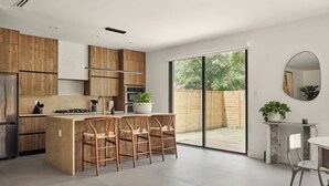 Una cocina moderna con armarios de madera, una isla central con taburetes de bar, un amplio ventanal corredizo a un patio trasero y un espejo redondo en la pared.