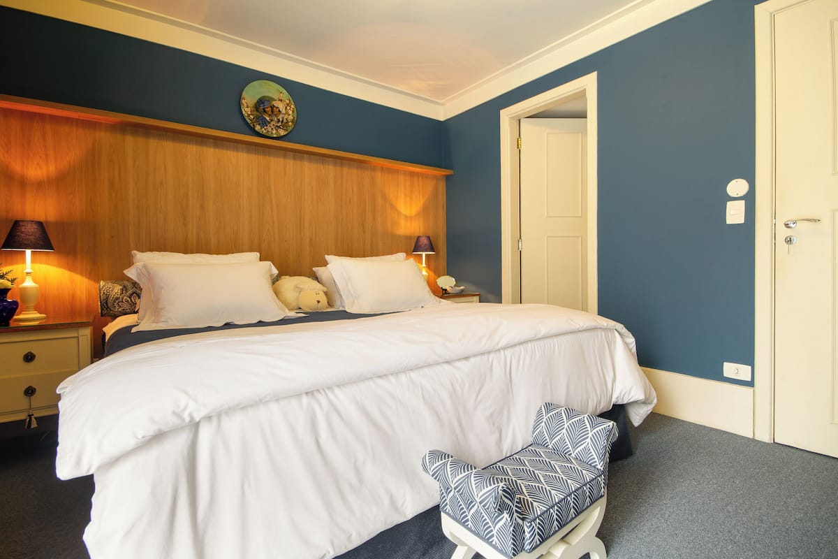 A neatly made bed with white linens, a bedside table with a lamp, and a bench with a patterned cushion.