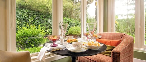 A dining area with a table set for tea, a wicker chair, and a view of lush greenery through large windows.