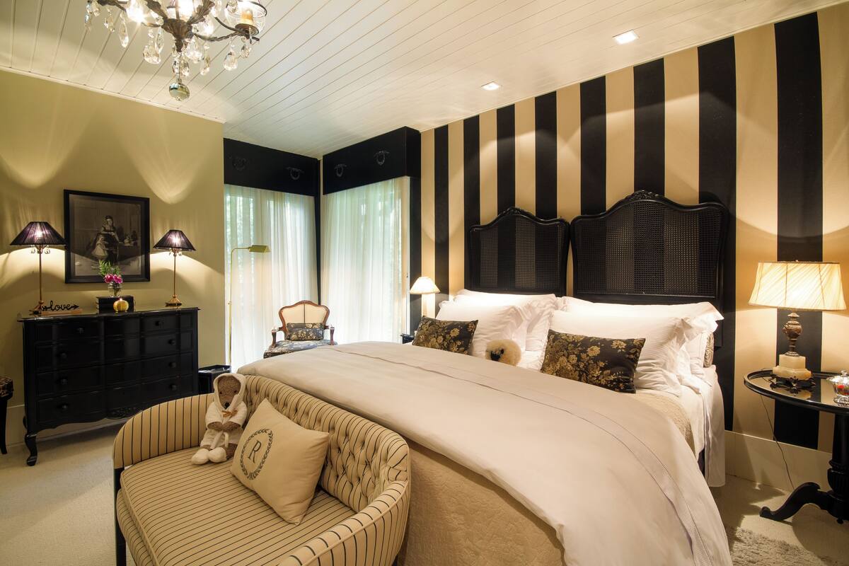 A hotel room with a large bed, a sitting area with a chair and a teddy bear, a dresser, a nightstand with a lamp, and a chandelier.