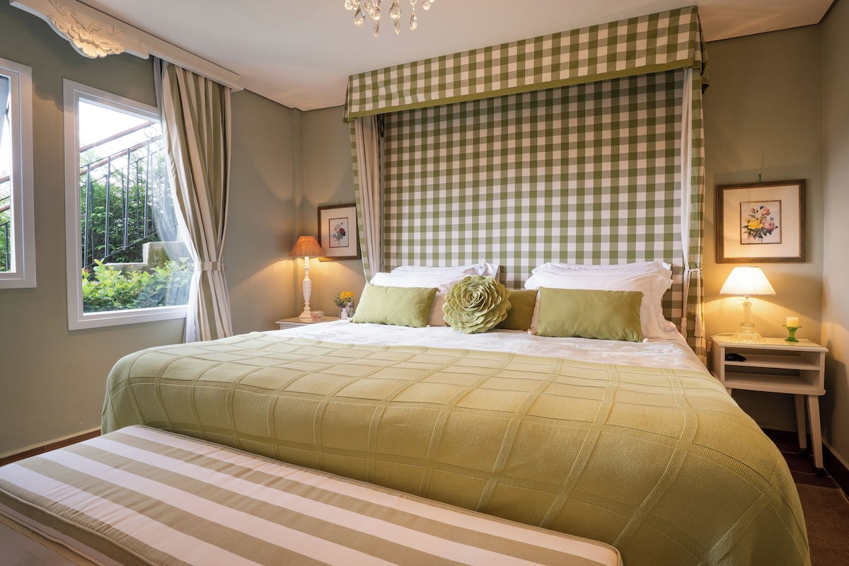 A neatly made bed with a green quilt, a bedside table with a lamp, and a window with curtains.