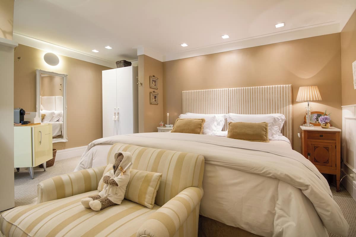 A bedroom with a large bed, a bedside table with a lamp, a dresser, and a chair with a teddy bear.
