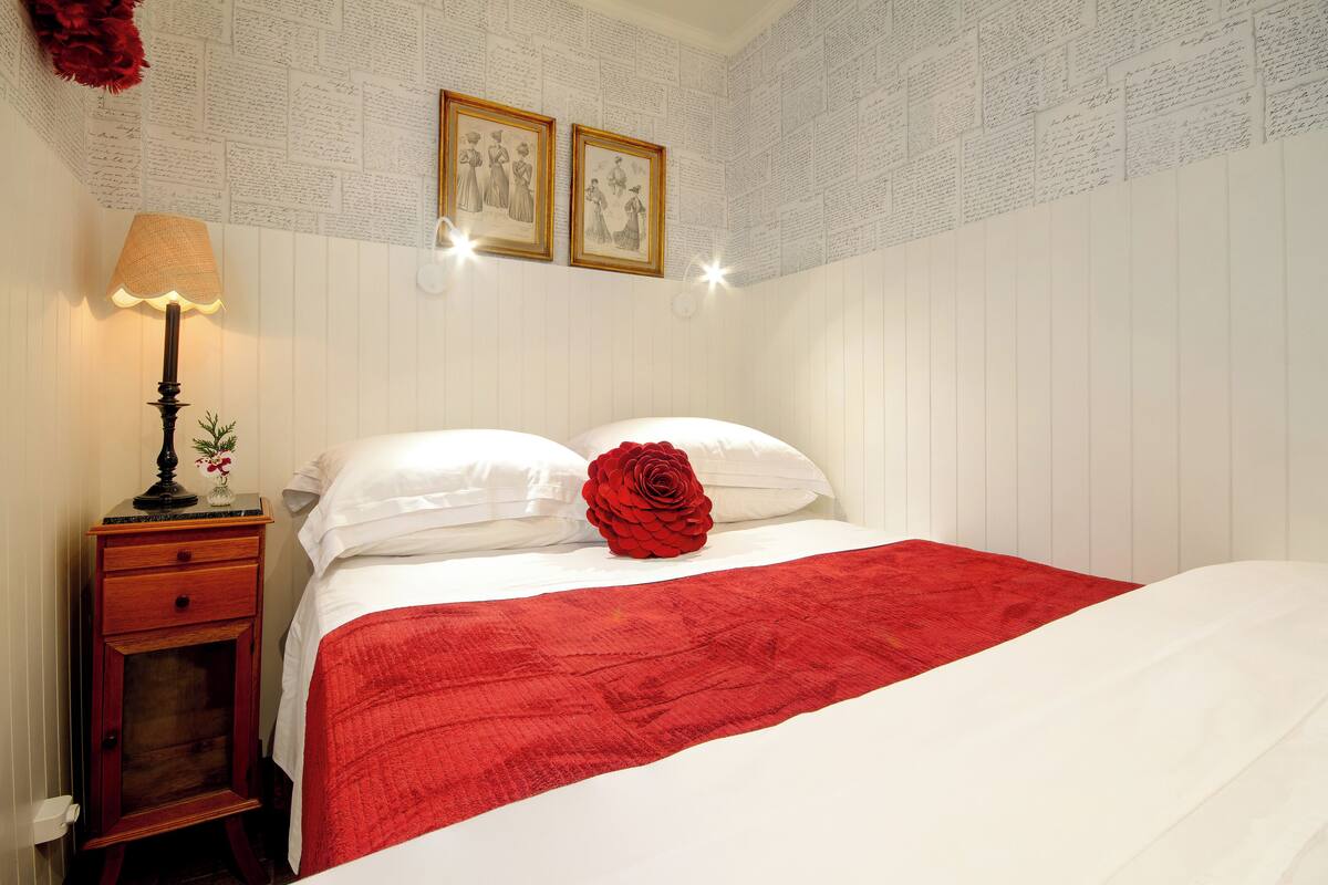 A neatly made bed with a red rose, white pillows, and a red blanket. There is a nightstand with a lamp and a small vase with flowers. Two framed pictures hang on the wall above the bed.