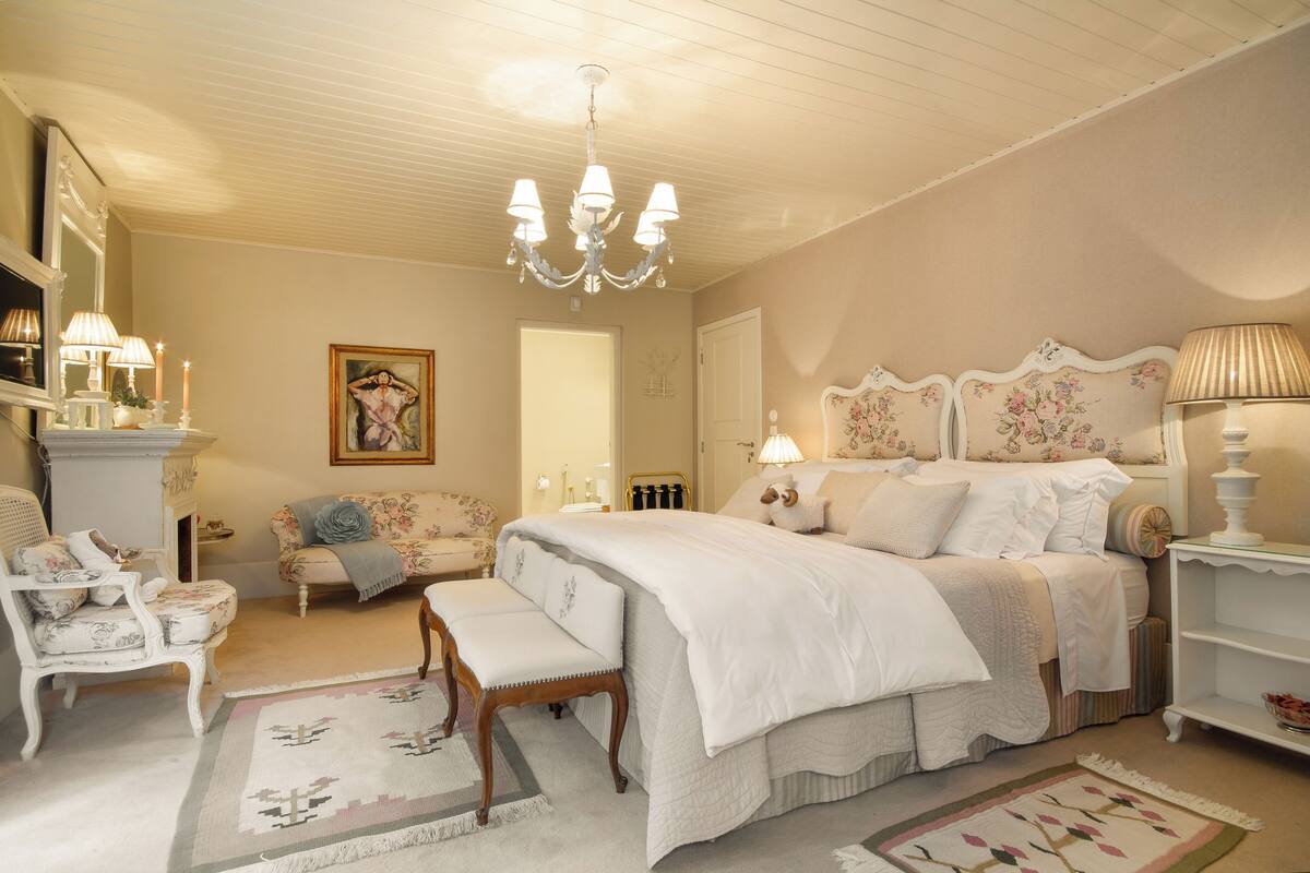 A bedroom with a large bed, a bench, a chair, a dresser, and a chandelier.